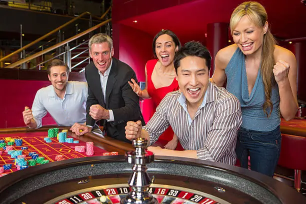 a group of joyful people at a roulette table in a casino, with several of them cheering and smiling, likely celebrating a win. The setting is vibrant with colorful gaming chips spread out on the table, and the atmosphere suggests excitement and entertainment.